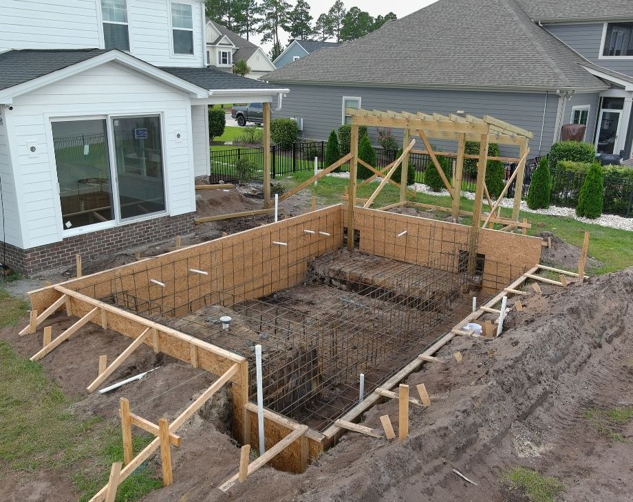 Construction site in a backyard showing early stages of a pool build, with wooden framing and rebar by a custom pool builder.