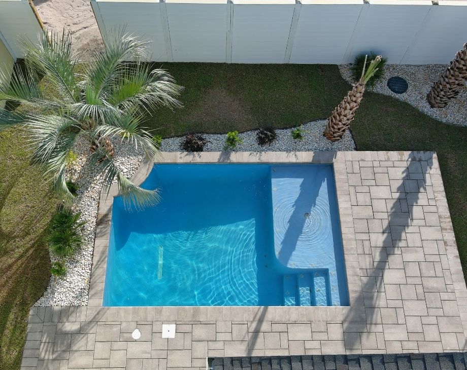 Small custom pool with stone patio surrounded by palm trees and white privacy fence, viewed from above.