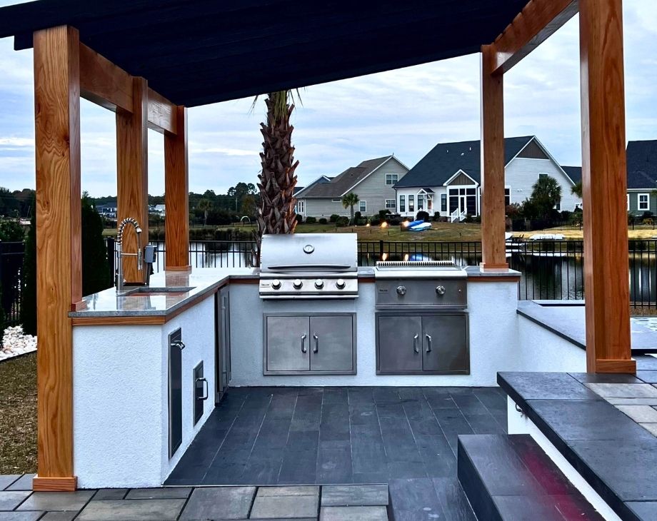 Outdoor kitchen with stainless steel grill under wooden pergola by a lake, ideal for a custom pool builder project.