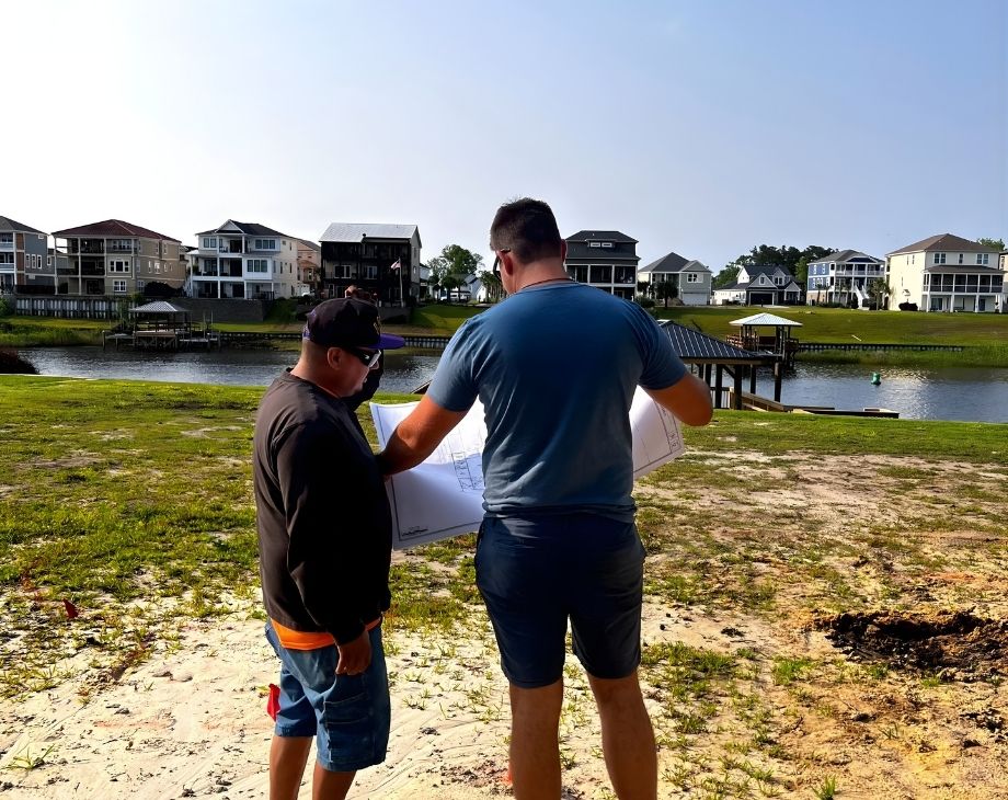 Two men examine blueprints near a lakeside. Residential homes visible, suggesting planning by a custom pool builder.