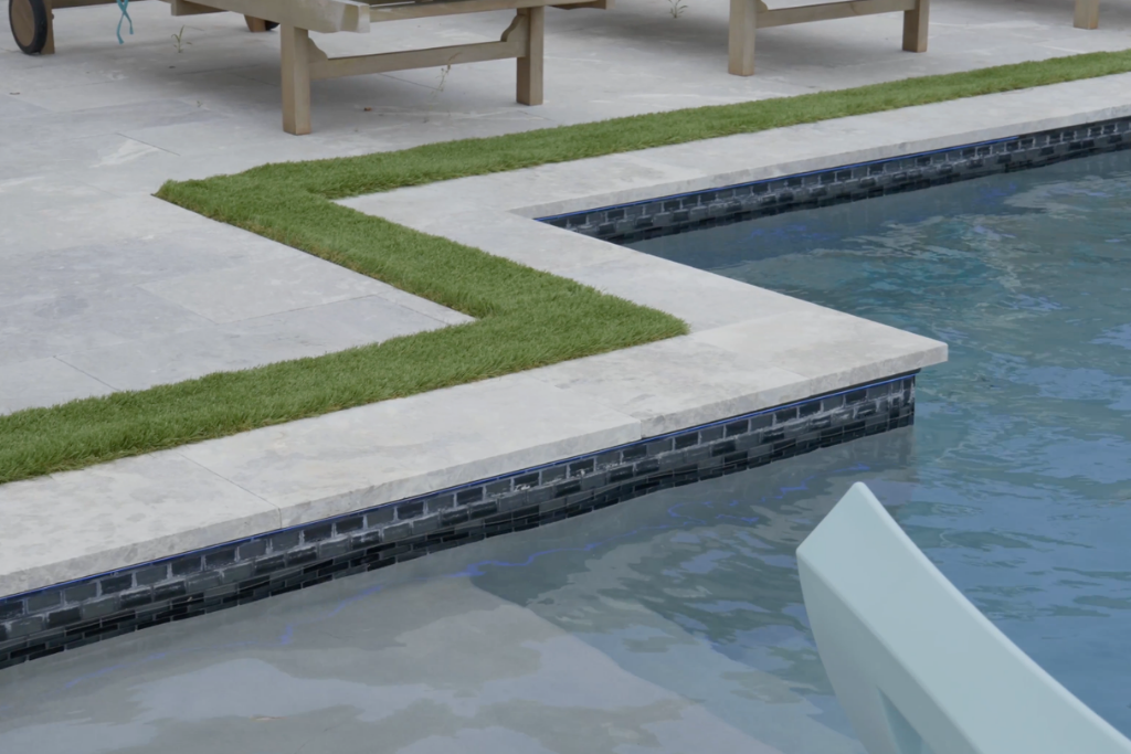 Elegant pool with angular design, surrounded by sleek stone tiles and grassy accents. Perfectly crafted by a custom pool builder.