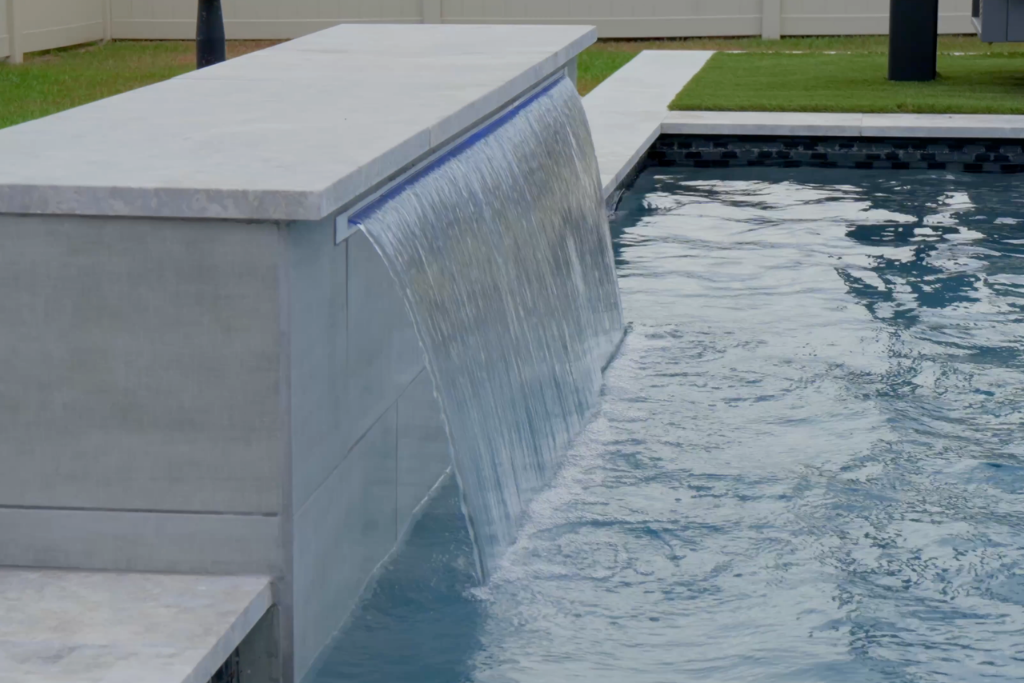 Elegant custom pool with a modern waterfall feature cascading into clear blue water, surrounded by a concrete border.