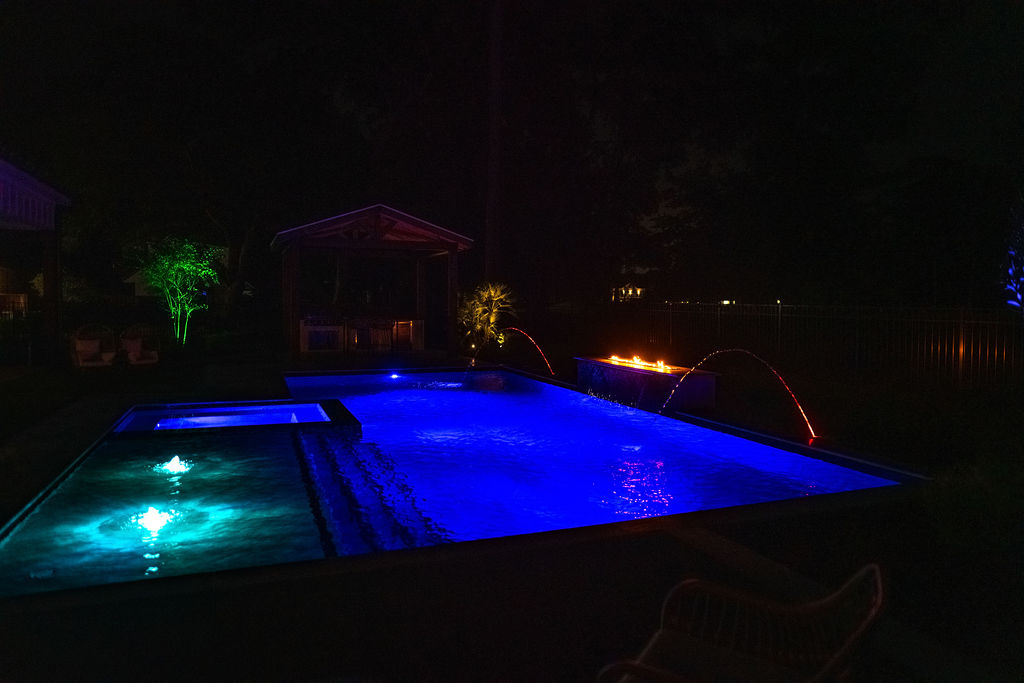 Illuminated custom pool with vibrant blue lights, fountains, and fire features, adjacent to a lit pavilion at night.