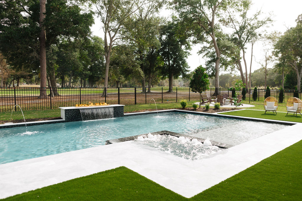 Modern backyard with a custom pool, featuring water fountains, a fire feature, and surrounded by lush greenery and seating areas.
