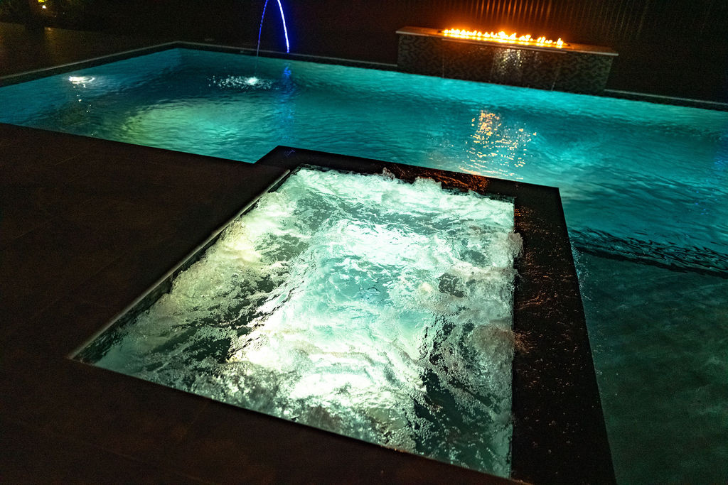 Luxurious night pool with spa jets and fire feature, showcasing custom pool builder's craftsmanship in elegant, illuminated design.