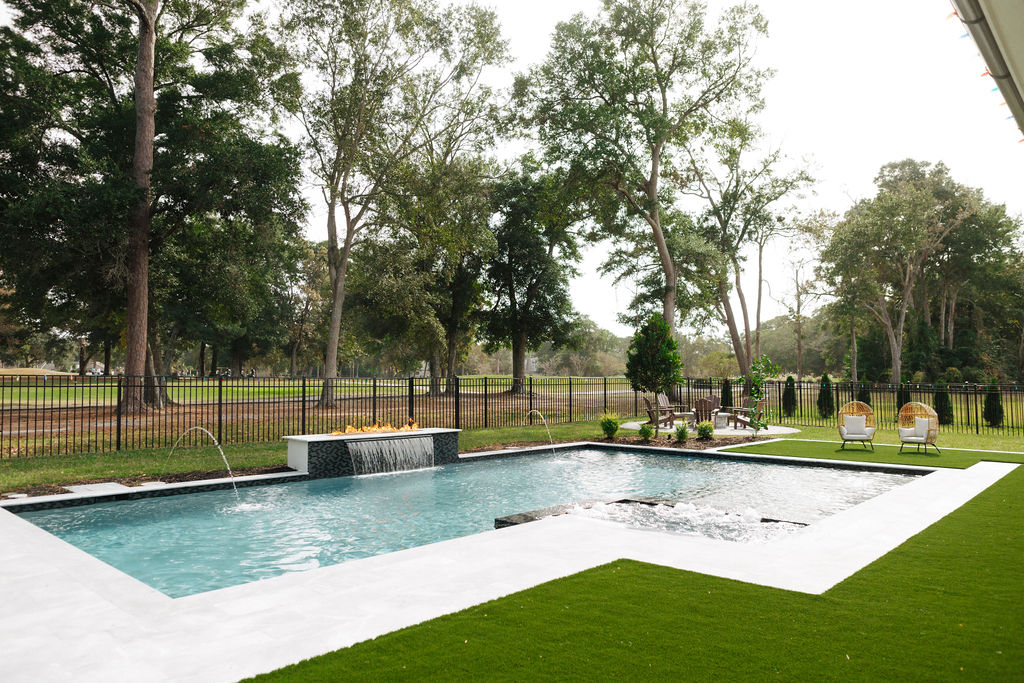 Modern custom pool with fountain and fire feature, surrounded by lush lawn and trees, bordered by a black fence and outdoor seating.