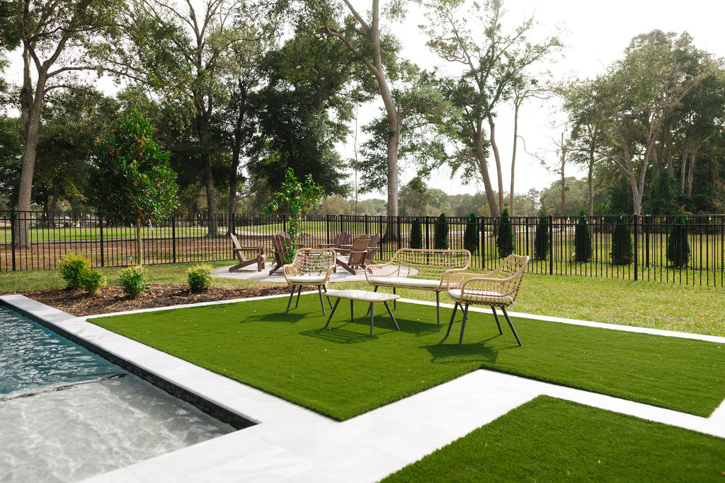 Modern backyard with a sleek custom pool, manicured lawn, wicker seating, and a backdrop of trees and a black metal fence.