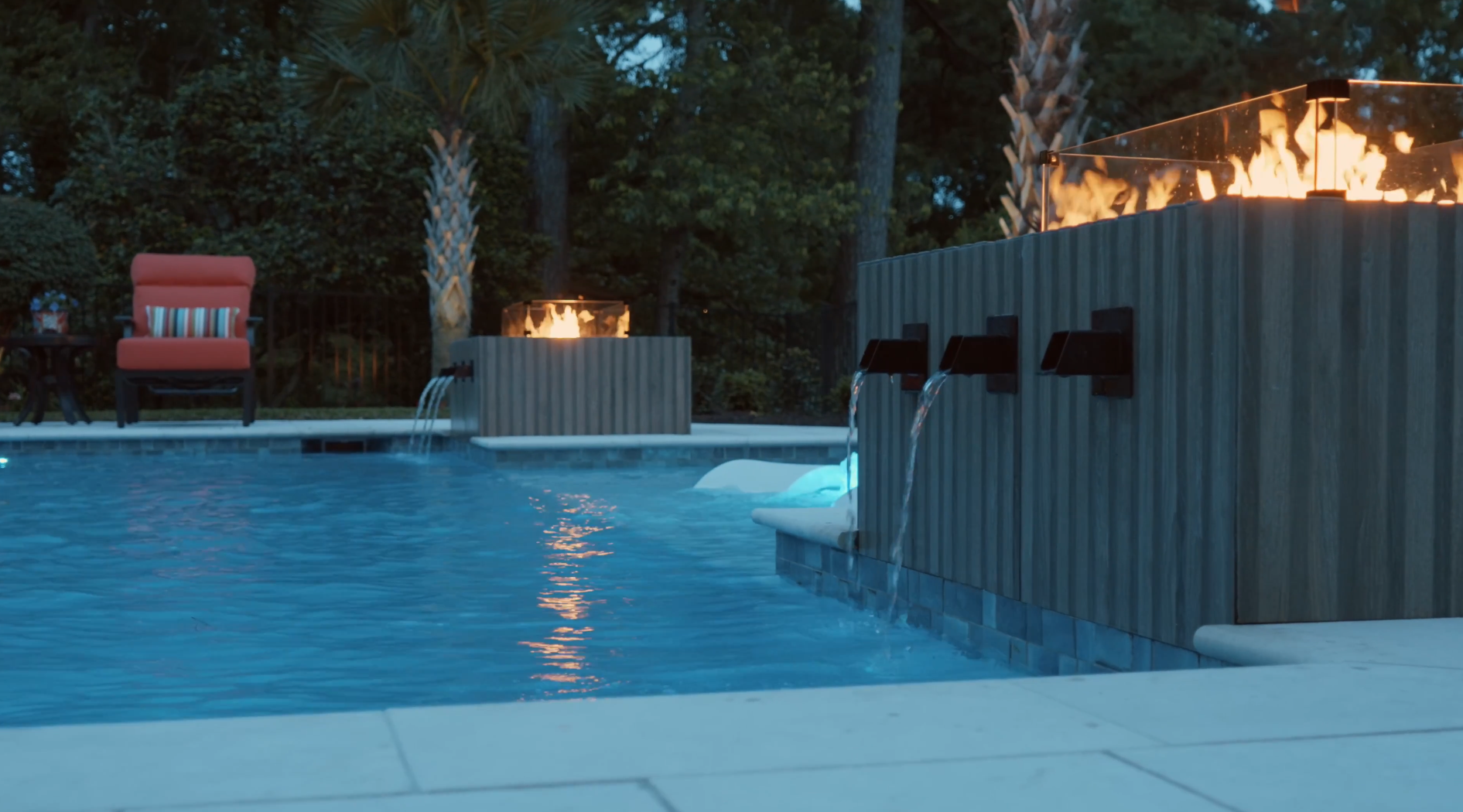 Luxurious pool with water features and fire accents, designed by a custom pool builder. Red chair and striped pillow nearby.