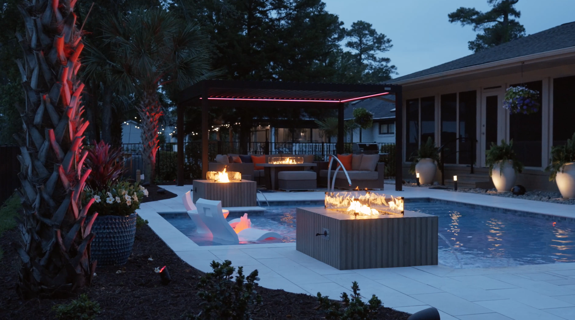 Modern backyard with pool by custom pool builder, featuring fire pits, pergola, palm trees, and evening lighting for a cozy outdoor ambiance.