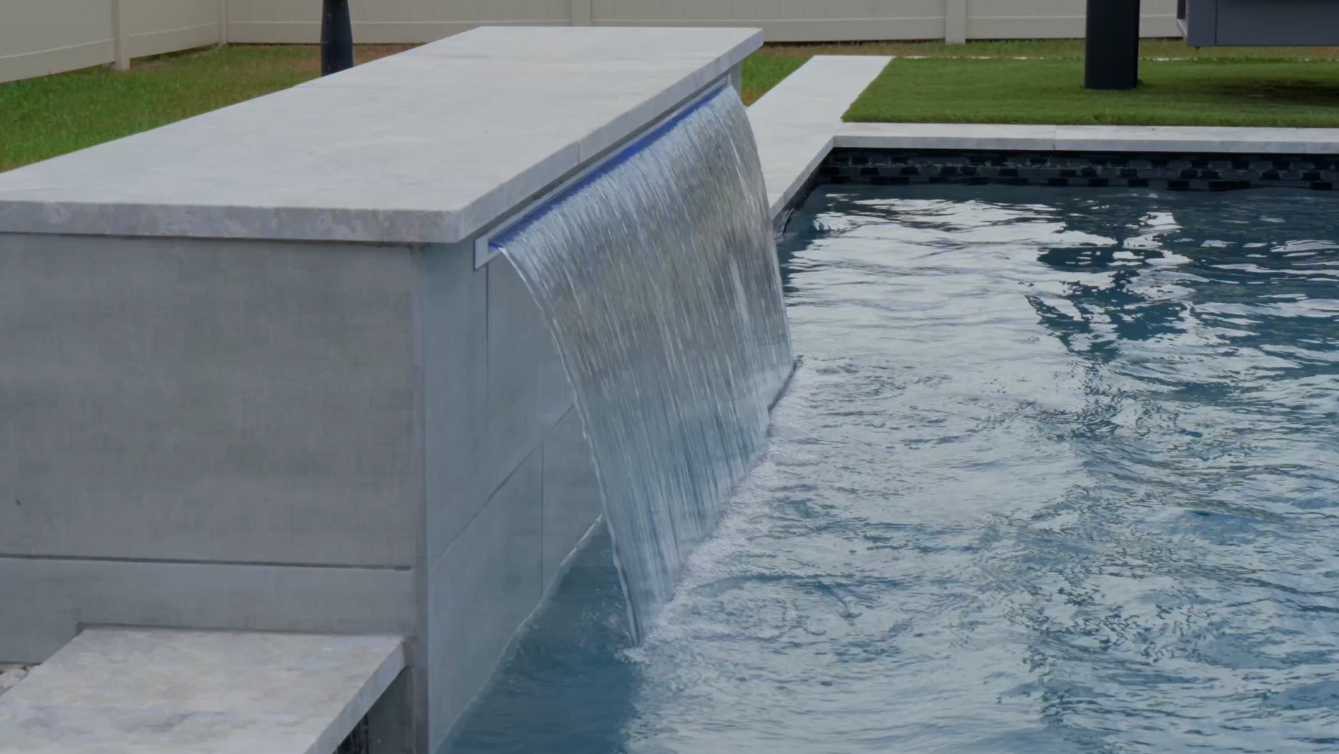 Water cascades from a sleek, modern pool ledge, demonstrating custom pool builder craftsmanship in a backyard setting.