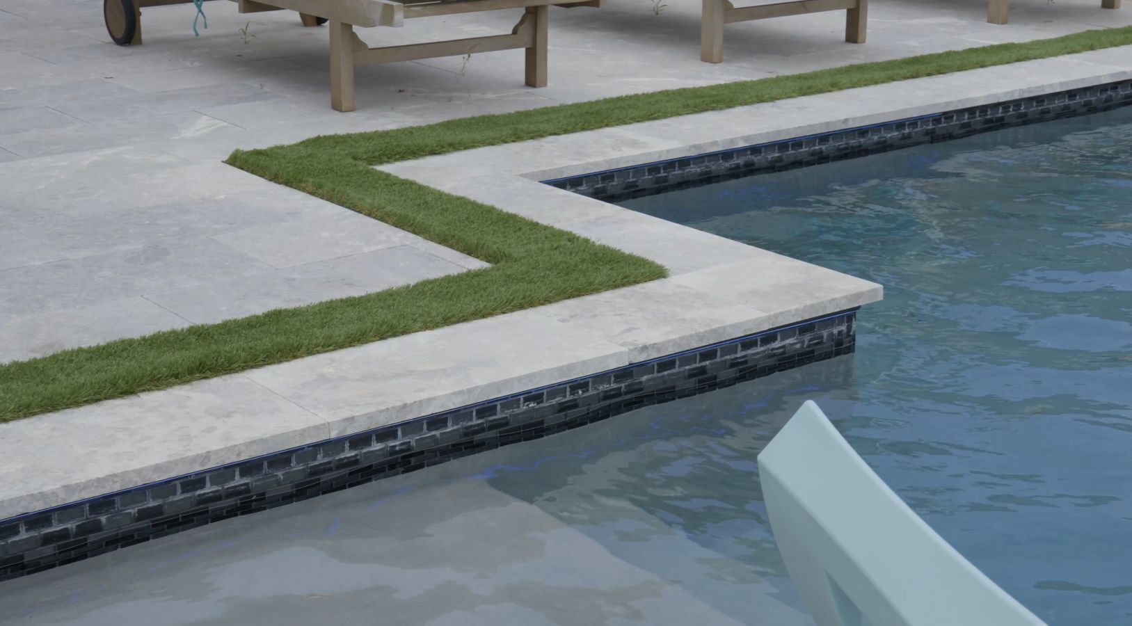 Elegant poolside area with grass borders, stone tiles, and clear blue water, showcasing expertise of a custom pool builder.