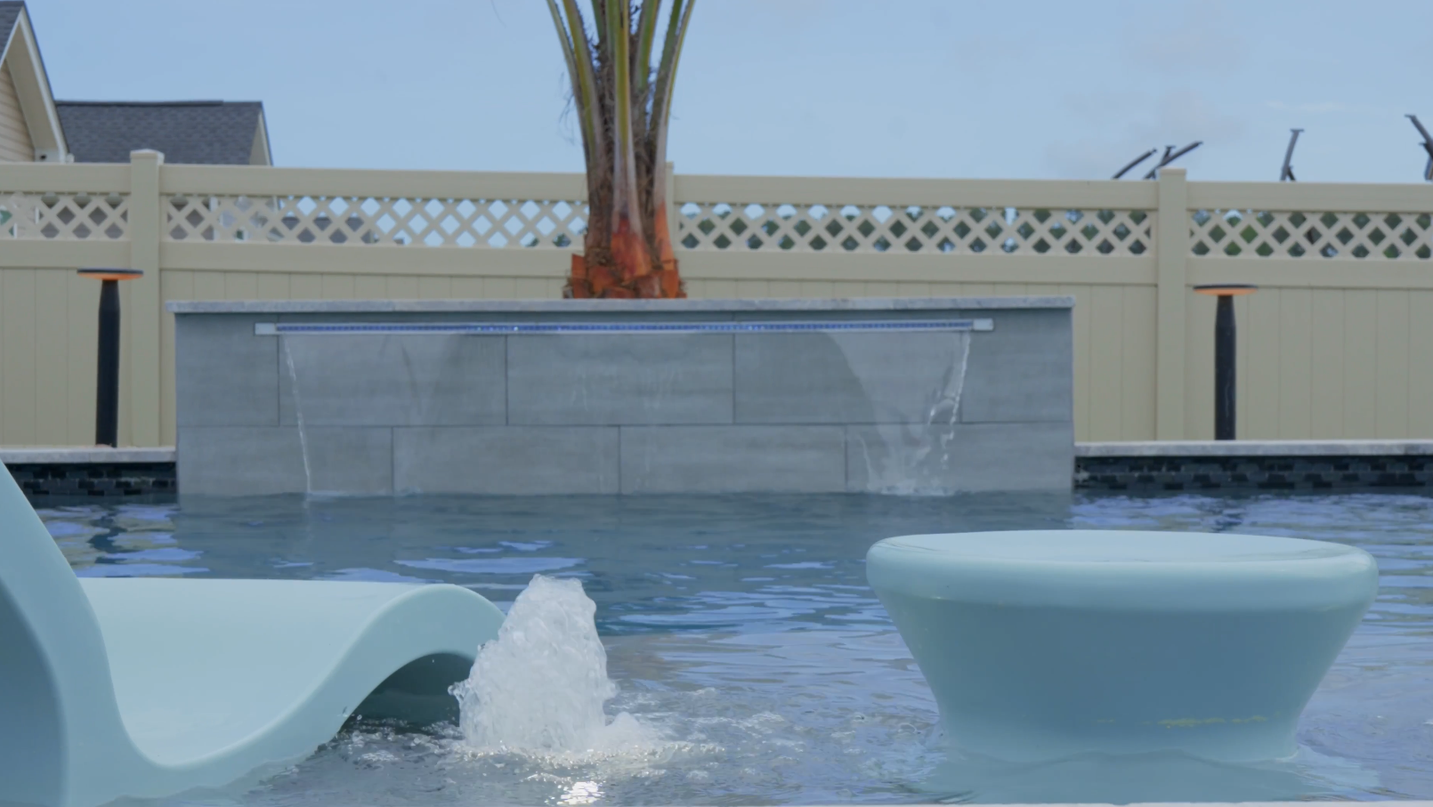 Modern pool with elegant fountains and sleek chairs, showcasing custom pool builder design. Lattice fence and tree in the background.
